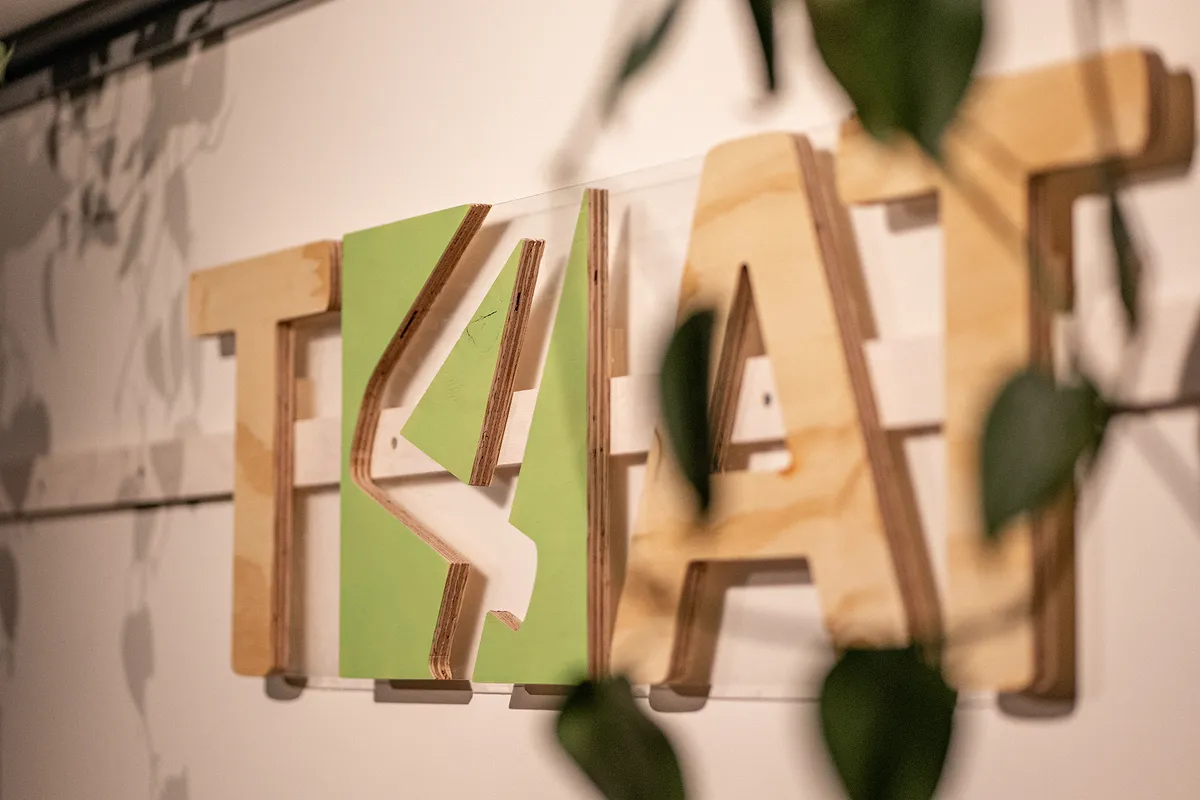 T4AT-Schriftzug aus Holz an der Wand, mit grünem Akzentbuchstaben und Pflanzenblättern im Vordergrund